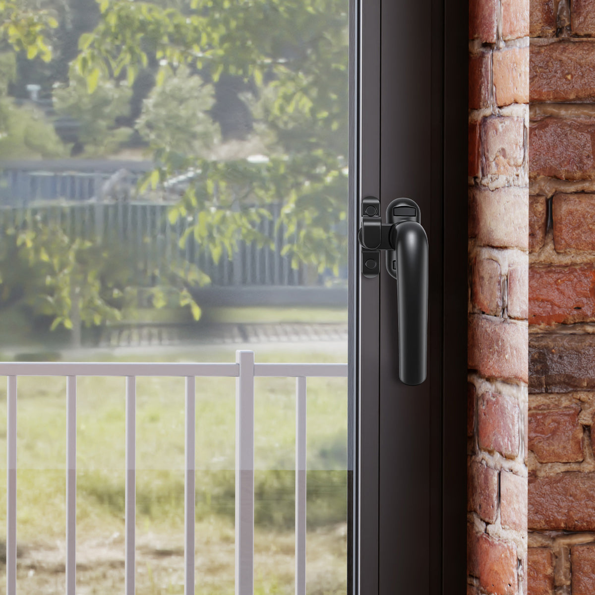 A close-up of a black metal window handle and latch mechanism on a dark-framed glass door, set next to a red brick wall with a blurred outdoor garden background visible through the glass.