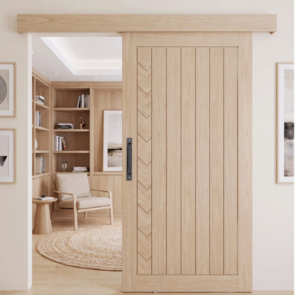 A light natural wood sliding door with vertical panels and a chevron inlay, shown installed in a modern home interior with a built-in bookshelf and chair visible through the opening.