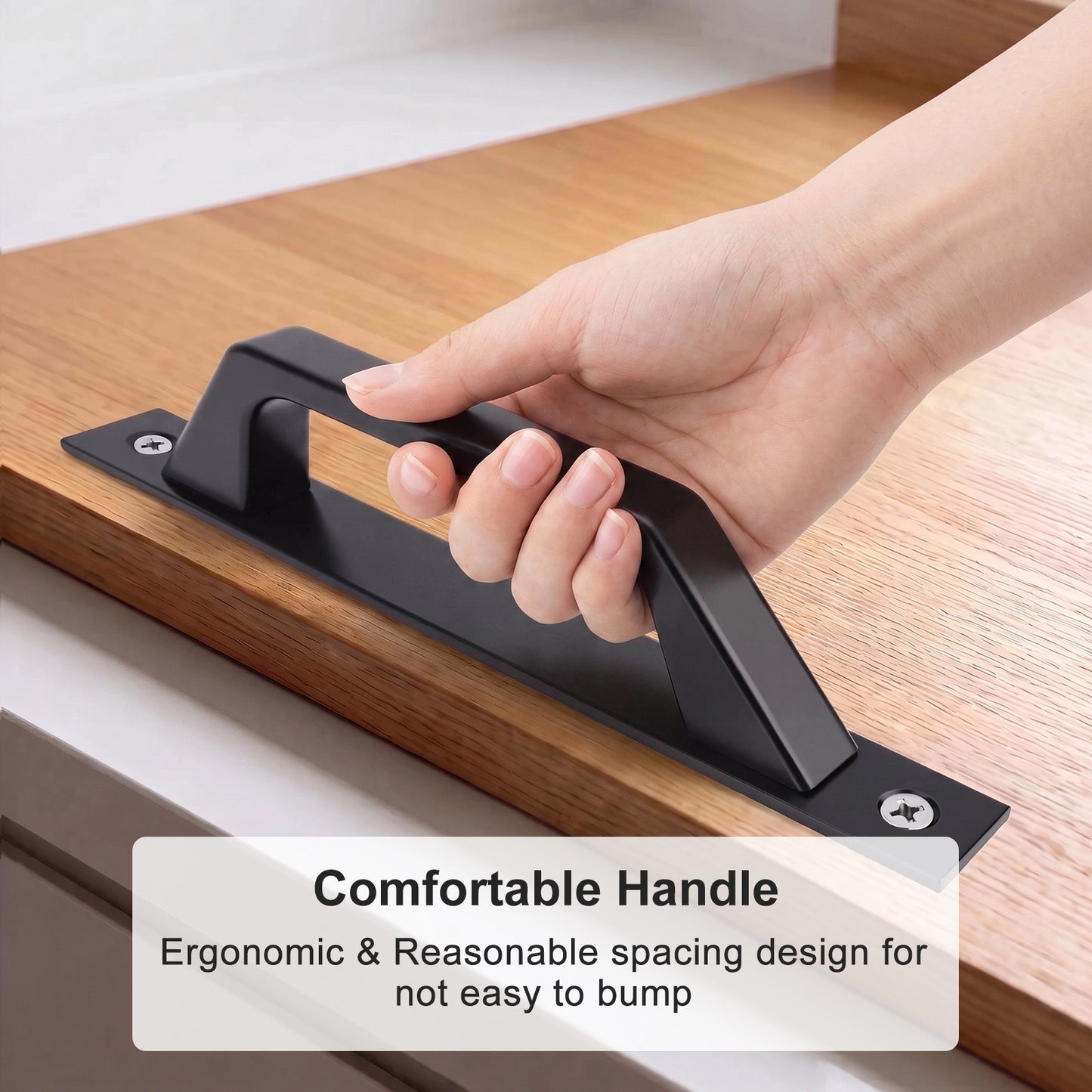 A hand holding a matte black cabinet pull handle mounted on a wooden drawer front, with text overlay reading 'Comfortable Handle' and 'Ergonomic & Reasonable spacing design for not easy to bump'.