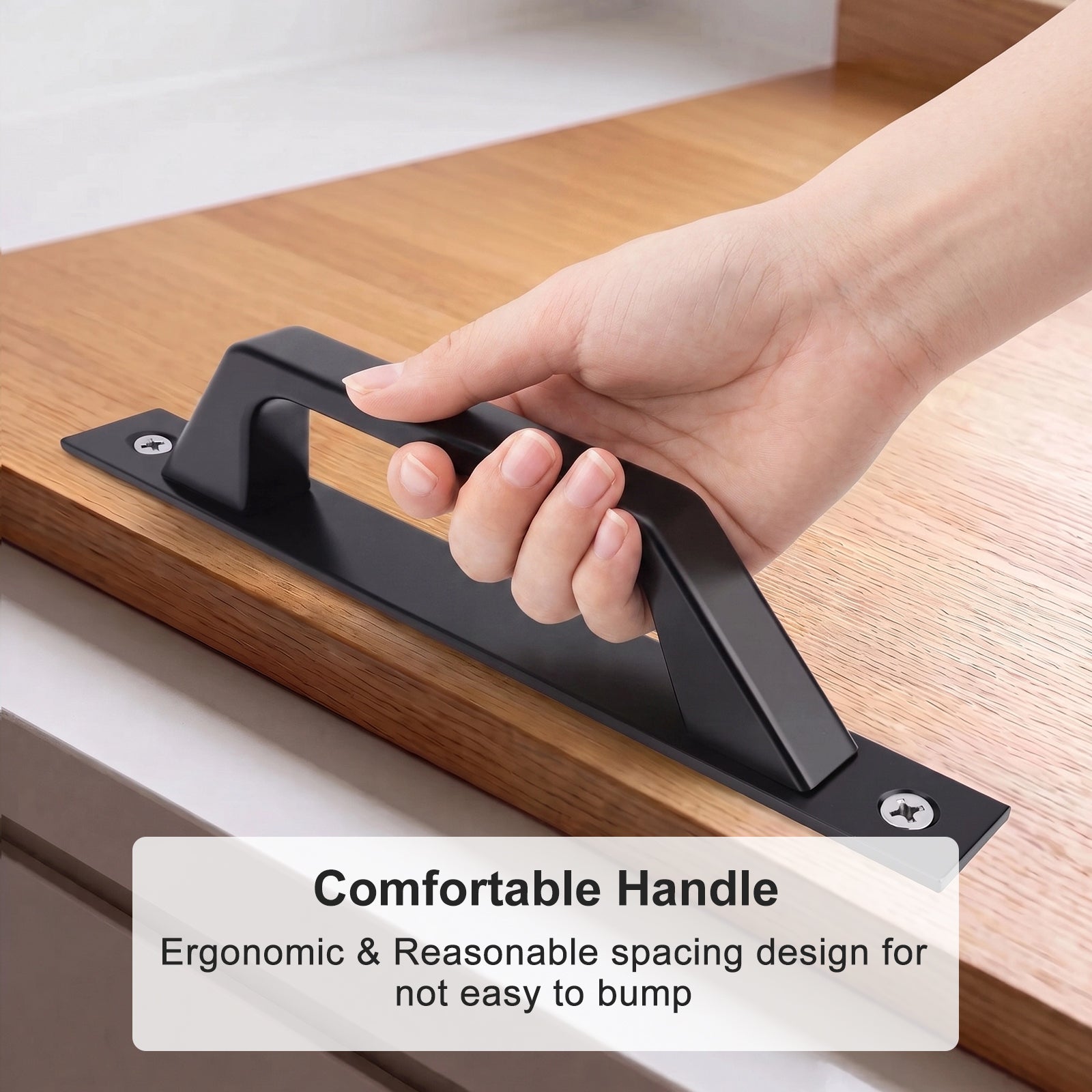 A hand holding a matte black cabinet pull handle mounted on a wooden drawer front, with text overlay reading 'Comfortable Handle' and 'Ergonomic & Reasonable spacing design for not easy to bump'.
